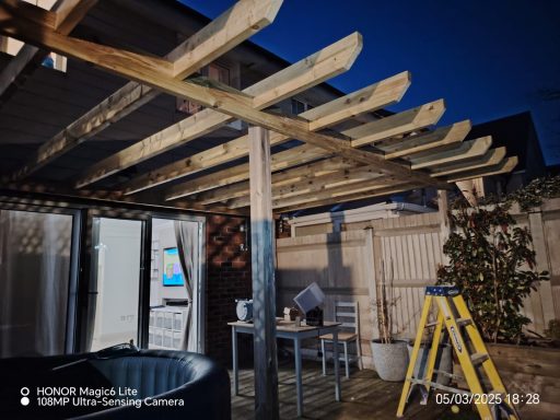 Wooden pergola in a backyard at night, with a hot tub and a ladder nearby.